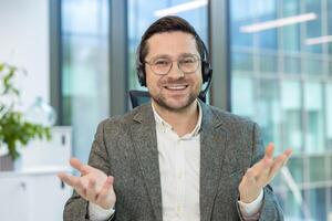 Portrait of a smiling young man in a headset sitting at an office desk and talking to the camera while gesturing with his hands. photo