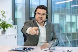 A man in a business suit, wearing a headset, points accusingly while looking directly at the camera. The office setting has natural light. photo