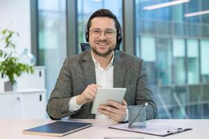 Portrait of a young man in a headset and glasses sitting at a working office desk in front of the camera, holding a tablet in his hands and looking at the camera with a smile. photo