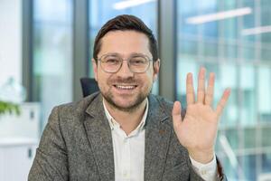 A smiling businessman in glasses waves to the camera, a friendly gesture captured in a professional setting. photo