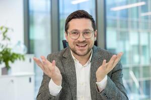 A young businessman in a suit and glasses sits in the office at a desk and talks to the camera while gesturing with his hands. photo