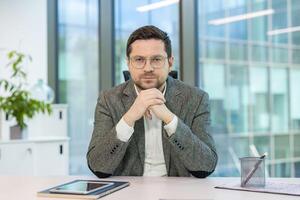 Portrait of a young successful and serious man in a suit and glasses sitting at a desk in the office, and looking confidently at the camera. photo