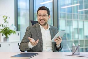A businessman in a conference, holding a tablet and gesturing in an office setting. He appears to be in a virtual meeting photo
