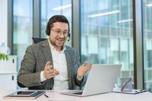 A businessman with glasses and headset participates in a conference, expressing himself with hand gestures while looking at the laptop screen. photo