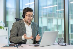 A young smiling man in a suit and headset is conducting a call on a laptop, selling, explaining and gesturing with his hands. photo
