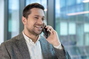 A smiling businessman in a grey blazer talks on his phone. The background is a modern office setting with glass windows. photo