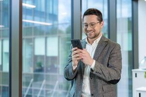 A smiling businessman wearing glasses, using a smartphone, standing near a large window in a modern office setting. photo
