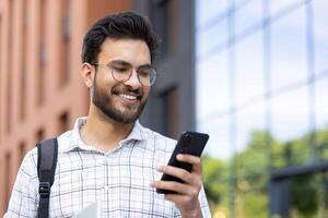 Young Indian man with beard and glasses standing outside on the street and smiling while using mobile phone. photo