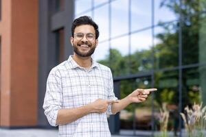 Portrait of a young Indian man in a shirt standing outside on the street, pointing with his fingers to the side at an empty space, smiling at the camera. photo