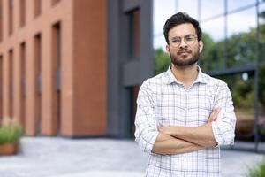 An Indian man with a serious expression stands in front of a modern building with his arms crossed. photo