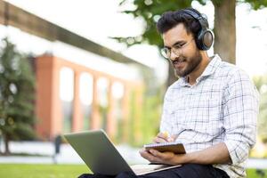 A young man, working on a laptop outdoors, wearing headphones and writing in a notebook. The background shows trees and a building. photo