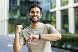 Portrait of an Indian young man standing outside, doing sports and running, looking at the camera with joy and satisfaction at the result on the watch timer on his wrist. photo