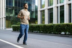 A fit, smiling man jogs down an urban street, enjoying a sunny day for his outdoor exercise routine. Modern city buildings are in the background. photo