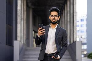 Portrait of young businessman outside office building, man smiling and looking at camera, holding phone, using app. photo