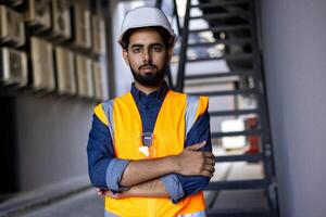 Serious engineer construction worker in helmet standing near building with crossed arms, arab work foreman looking and camera, senior project worker designer. photo