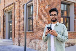 Portrait of young successful student outside university campus, man smiling looking at camera and using app on phone, typing message and browsing social media, Indian man holding phone with backpack. photo