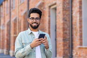 Casual arab male looking at camera with wide smile while using cellphone on brick building background. Careless gadget user in classic spectacles enjoying playing new online mobile application. photo