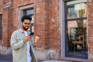 Young man walking in the city reading messages on the phone smiling, with a cup of hot drink, businessman entrepreneur programmer in casual clothes using an application on a smartphone. photo