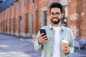 Relaxed middle eastern man holding telephone and plastic coffee cup while walking during daytime. Positive brunet freelancer with flexible work schedule taking break for beverage and scrolling feed. photo