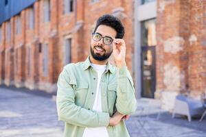 Interested indian man with beard touching eyeglasses and looking aside while passing brick building. Carefree young person having walk in old district while doing sightseeing and getting impressed. photo