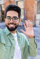 Phone web camera view, young man uses app on smartphone for remote call communication, Indian man smiles and looks at camera, waves hand in greeting. Vertical shot. photo