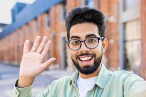 Phone web camera view, young man uses app on smartphone for remote call communication, Indian man smiles and looks at camera, waves hand in greeting. photo