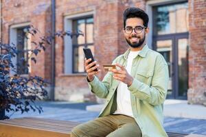 Portrait of a happy young Hindu man using a credit card and phone, sitting outside on a bench and smiling at the camera. photo