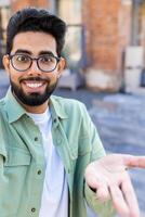 Phone web camera view, young man uses app on smartphone for remote call communication, Indian man smiles and looks at camera, waves hand in greeting. Vertical shot photo