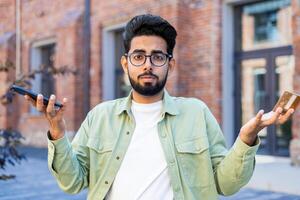 Portrait of uncertain man in eyewear spreading hands in confusion while holding modern smartphone outside. Unhappy male feeling displeased with loosing connection on gadget with technical problems. photo