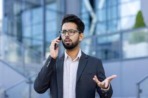 Close-up photo of an angry and sad young Indian man in a business suit standing on the street near an office center and talking ,emotionally on the phone while gesturing with his hands.