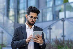 A professional Indian businessman, wearing glasses, is intently using a tablet outside modern glass buildings. The concept of mobile business communication in a global setting is depicted. photo