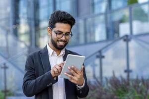 Close-up photo of a smiling young Indian man in a suit sitting near an office building and using a tablet.