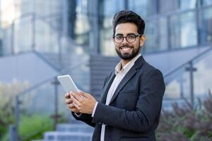 A cheerful Indian businessman in formal attire stands outdoors, using a digital tablet with a confident smile. Modern business setting with a multicultural subject. photo