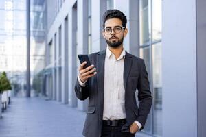 A professional Indian businessman in a smart suit stands outside modern office buildings, intently using his smartphone. This image conveys a sense of connectivity, confidence, and the fast pace of corporate life. photo