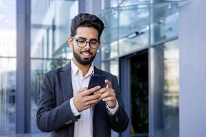 A young Indian businessman in a suit is engaged with his smartphone, smiling as he texts or reads a message. This image captures a moment of modern professional life, possibly during a break or while commuting. photo