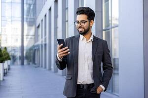A happy and successful young Indian man in formal wear is engaged with his smartphone, smiling while standing in a modern city environment. This image captures the essence of connectivity, urban life, and professional success. photo