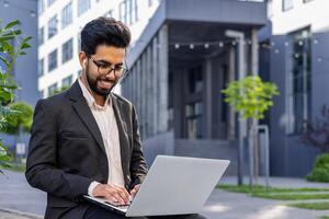 A smartly-dressed professional man engages with a laptop while seated outside a contemporary office setting, embodying productivity and determination in an urban environment. photo