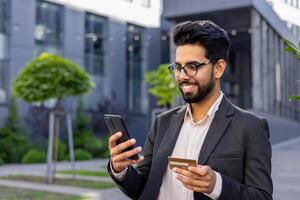 A cheerful Indian man stands outside an office complex, expertly handling a smartphone in one hand and a credit card in the other. The image exudes secure financial transactions. photo