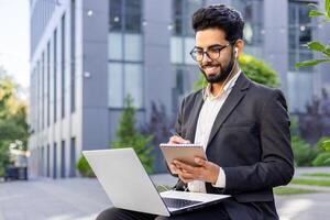 A focused young adult sitting outside a modern building, working intently on a laptop while taking notes. Projects the concept of remote work, busy professionals, and urban workspaces. photo