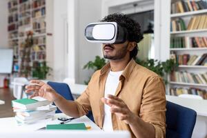 A young Indian male student is sitting in a library and studying through a virtual mask online. Sitting at the table and gesturing with his hands. Close up photo. photo