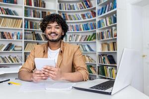 Close-up photo of a young Indian male student working in a university library, sitting at a desk, using a tablet and smiling at the camera.