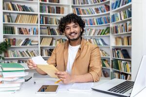 Portrait of a young Muslim male student sitting at a desk in a university library and opening a received letter, smiling and pointing at the camera. photo