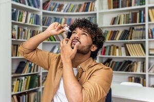 A young adult male is captured applying eye drops while standing in a cozy home library. The image reflects self-care and health management against a backdrop of bookshelves filled with books. photo