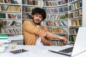 Young tired guy stretching hand and shoulder while looking at notebook on desk with books and computer. Bored male making light exercise for reducing muscle tension before going back to work. photo