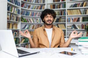 Middle eastern man sitting by desktop with computer and keeping raised hands in gyan mudra with closed eyes. Calm programmer using yoga pose practising for getting rid of stress and freeing mind. photo