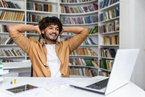 Positive hispanic guy leaning on back of chair while holding hands behind head and looking at laptop screen. Smiling student feeling satisfied after passing exam remotely and getting high grade. photo