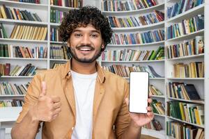 A young man stands in front of a bookshelf in a library, presenting a smartphone with a blank screen, giving a thumbs-up sign, suggesting approval or a positive review of an app. photo