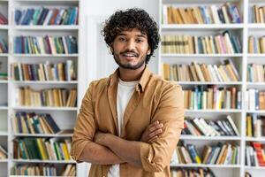 Confident diverse man in casual outfit looking at camera with arms folded on chest on background of closet with books. Intelligent male posing in library of university campus with happy expression. photo