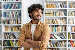 A casually dressed professional stands assuredly with arms crossed in front of a bookshelf. The focus is on confidence and casual professionalism within an educational or literary setting. photo