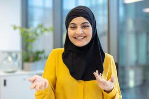 Smiling woman in hijab gestures, engaged in a call, looking directly at the camera, seemingly talking to someone photo